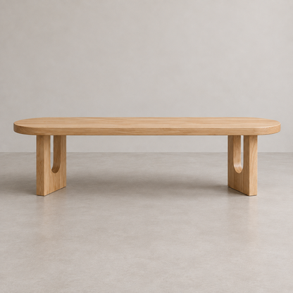 A minimalist light oak wood bench with a rounded rectangular top and two integrated block legs featuring circular cutouts, set against a neutral gray studio background.