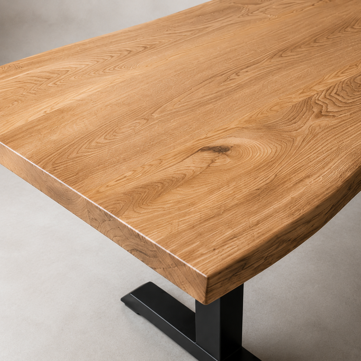 A close-up of a live-edge wooden dining table with a warm elm finish and a black metal pedestal base, shown on a light gray concrete floor.