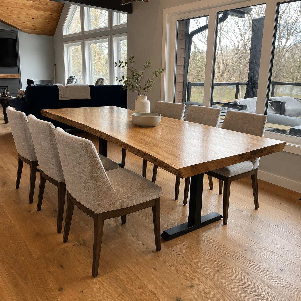 A modern dining room scene featuring a rectangular live-edge wooden table with black metal T-style legs, surrounded by six upholstered dining chairs with dark wood frames. The room has light oak flooring, large windows, and a dark sofa in the background.