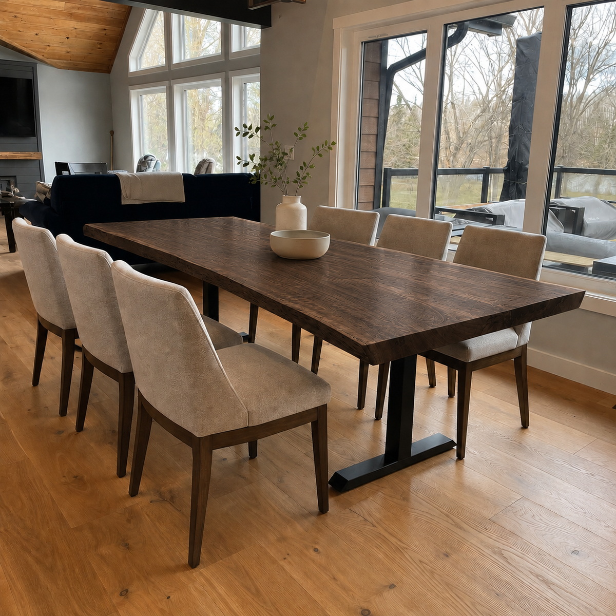 A dark wood dining table with black metal T-style legs and six upholstered dining chairs in a bright, modern dining room with hardwood floors and large windows.